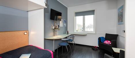 Ein kleines Hotelzimmer mit einem Bett, einem Schreibtisch, zwei Stühlen, einem Fenster und einem an der Wand montierten Fernseher.