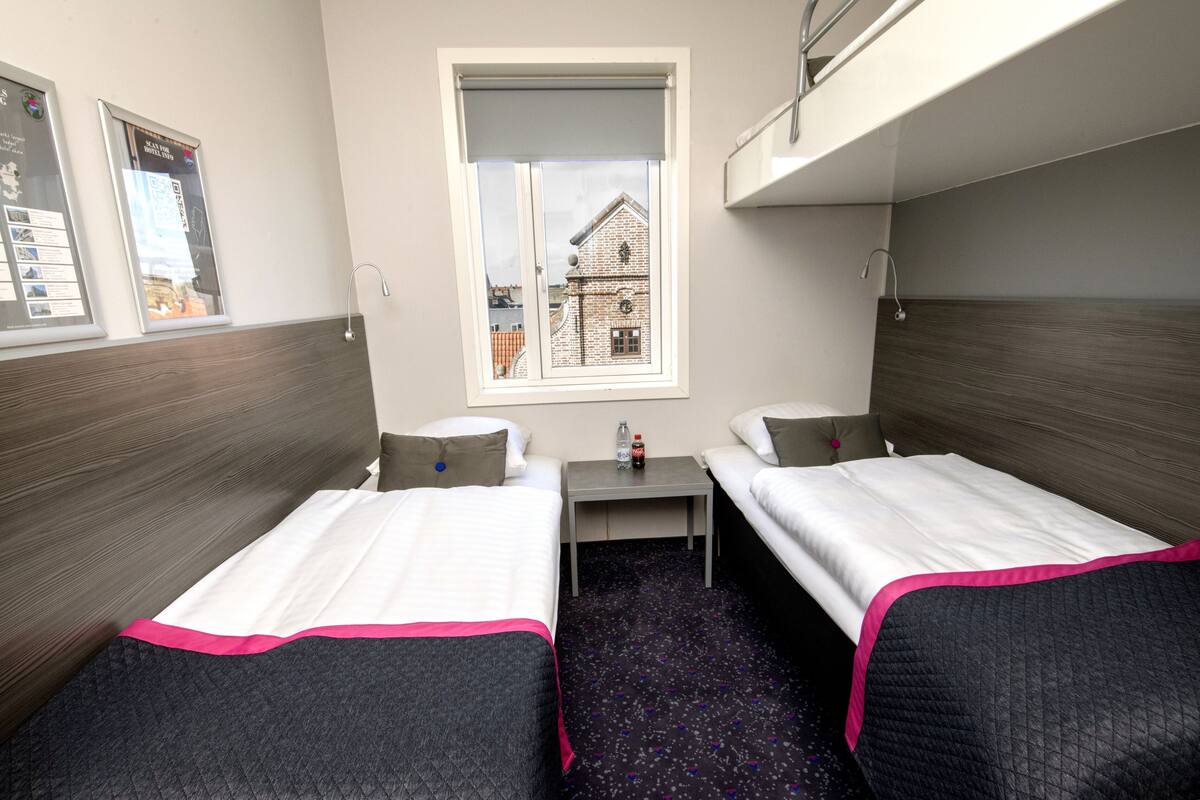 A compact hotel room with two single beds, a small table, a window with a view of a building, and a bunk bed above.