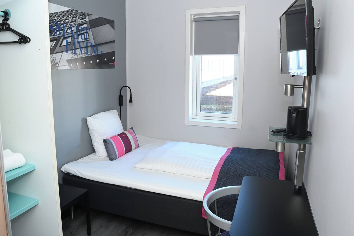 A compact hotel room with a bed, a desk, a chair, a window with a view, a wall-mounted TV, and a coat rack.