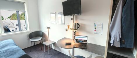 A hotel room with a bed, a desk with a laptop, a chair, a TV mounted on the wall, and a wardrobe.