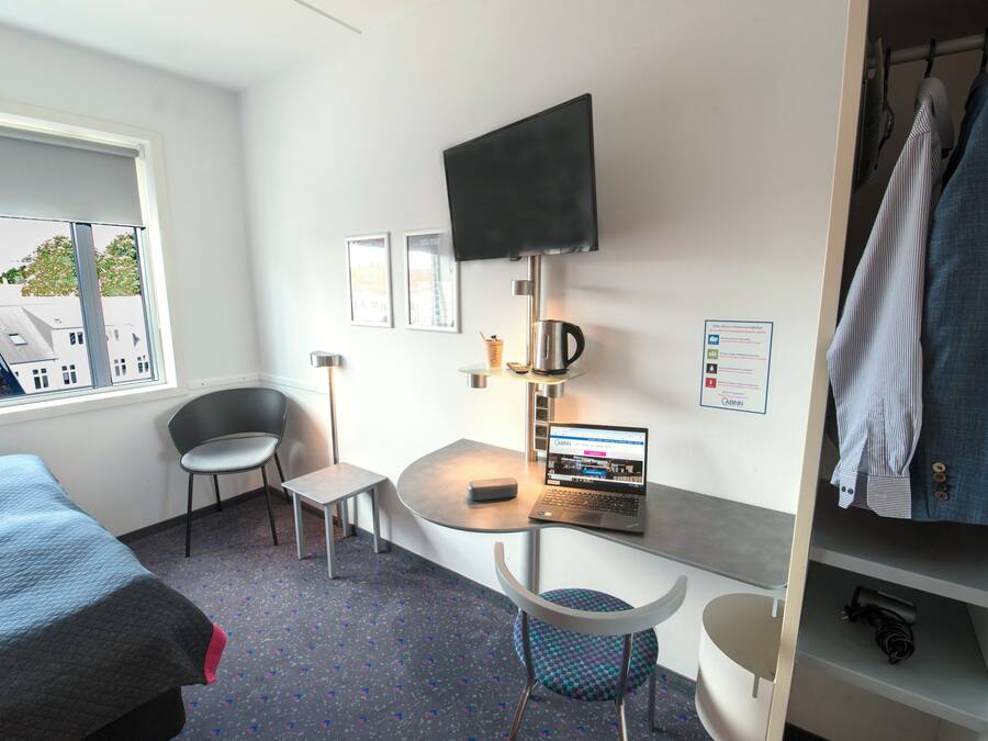 Habitación de hotel con cama, escritorio con una computadora portátil, una silla, televisor montado en la pared y un armario.
