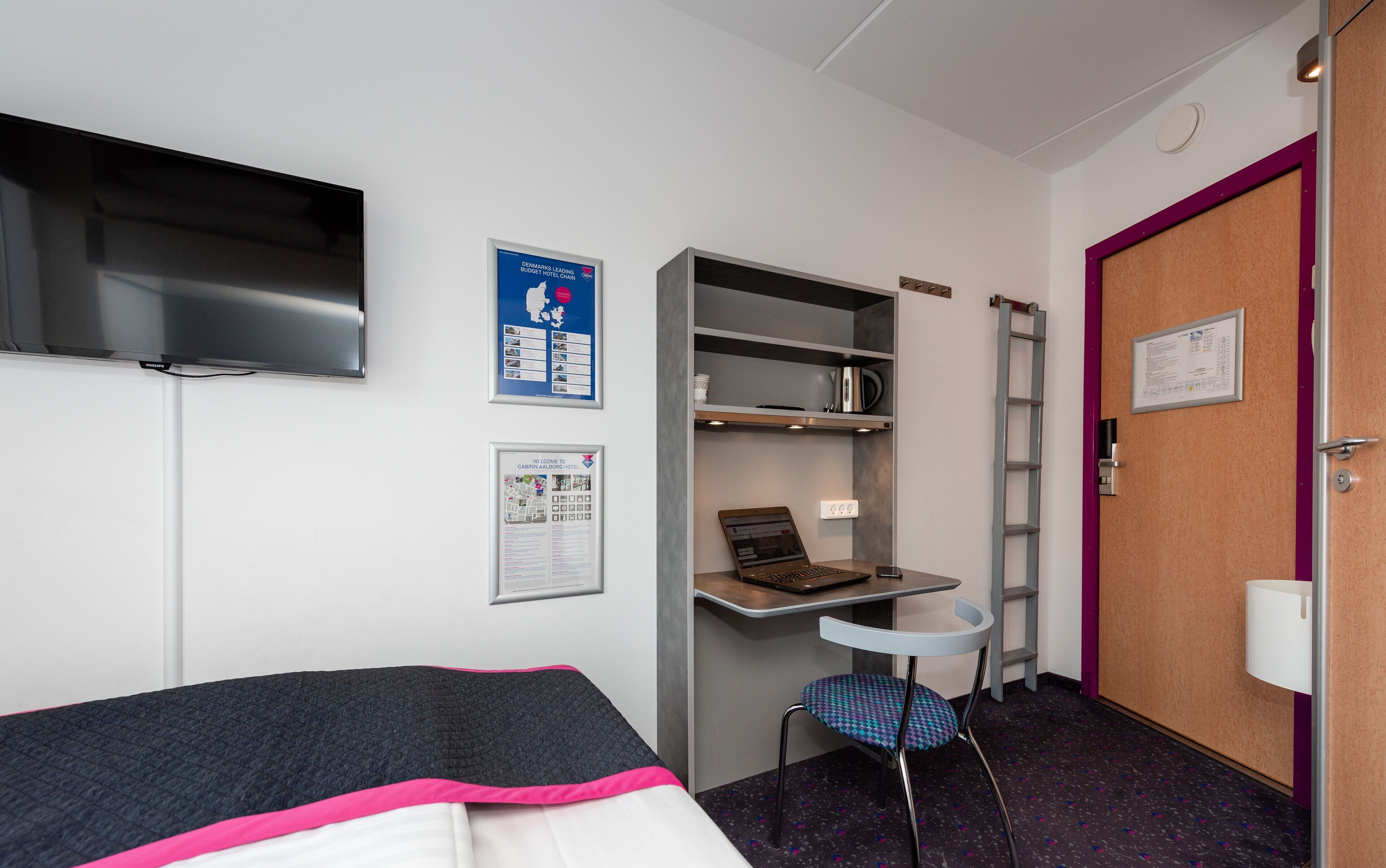 A compact hotel room with a bed, a desk with a laptop, a TV, and a small chair.