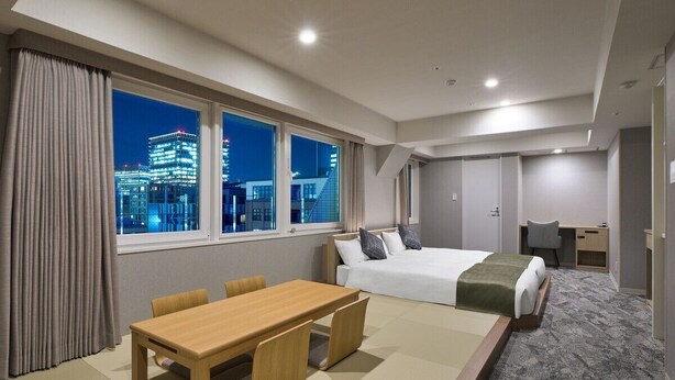 Ein Hotelzimmer mit Bett, Schreibtisch, Stuhl, Tisch und Blick auf eine nächtliche Stadtansicht.