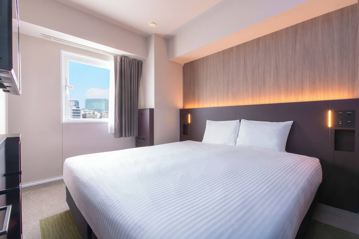 A hotel room with a large bed, a window with curtains, a wall-mounted TV, and a wall-mounted light.
