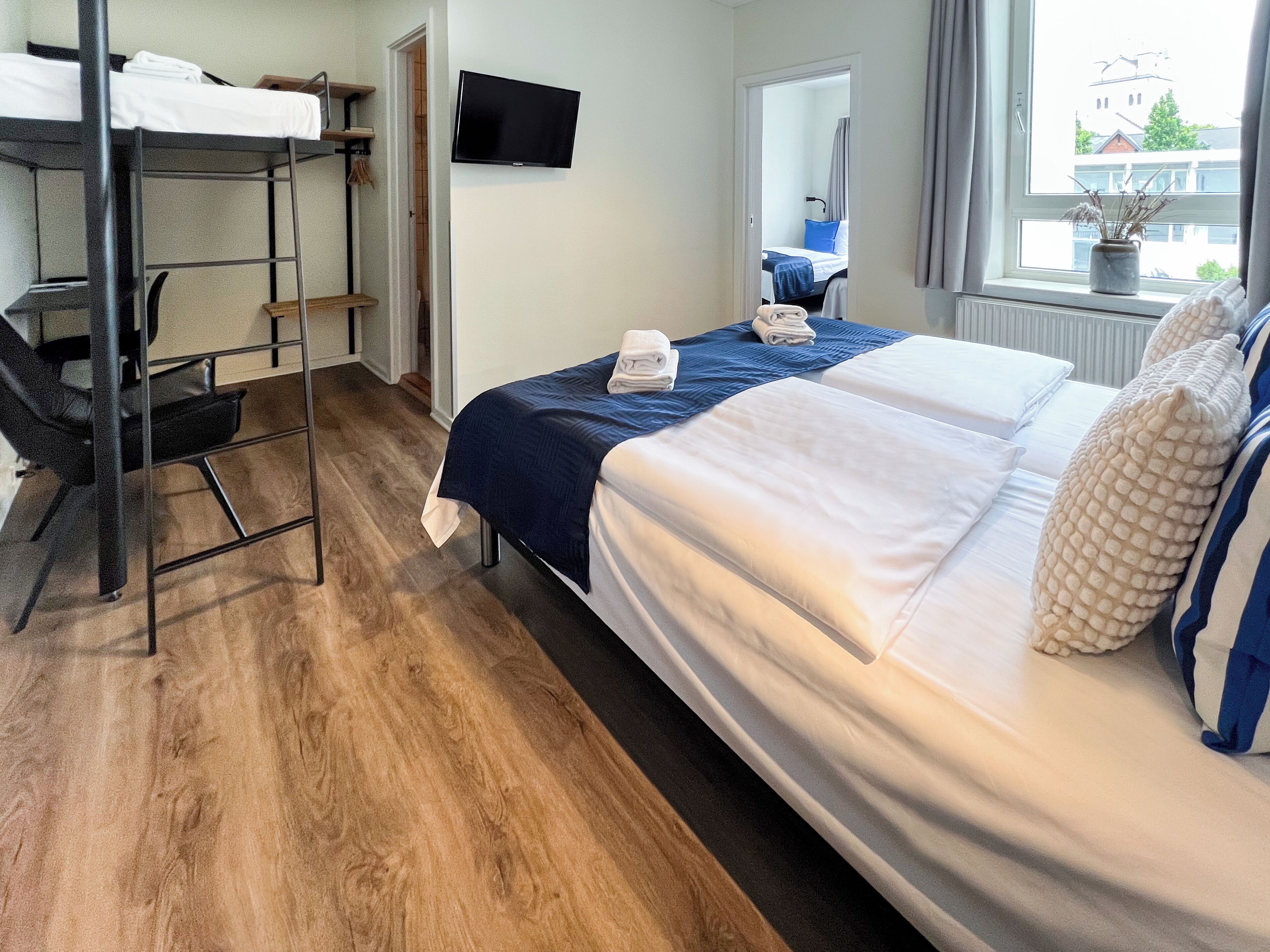 Une chambre d’hôtel avec un lit superposé, un bureau, une chaise, une télévision et une fenêtre donnant sur des immeubles.