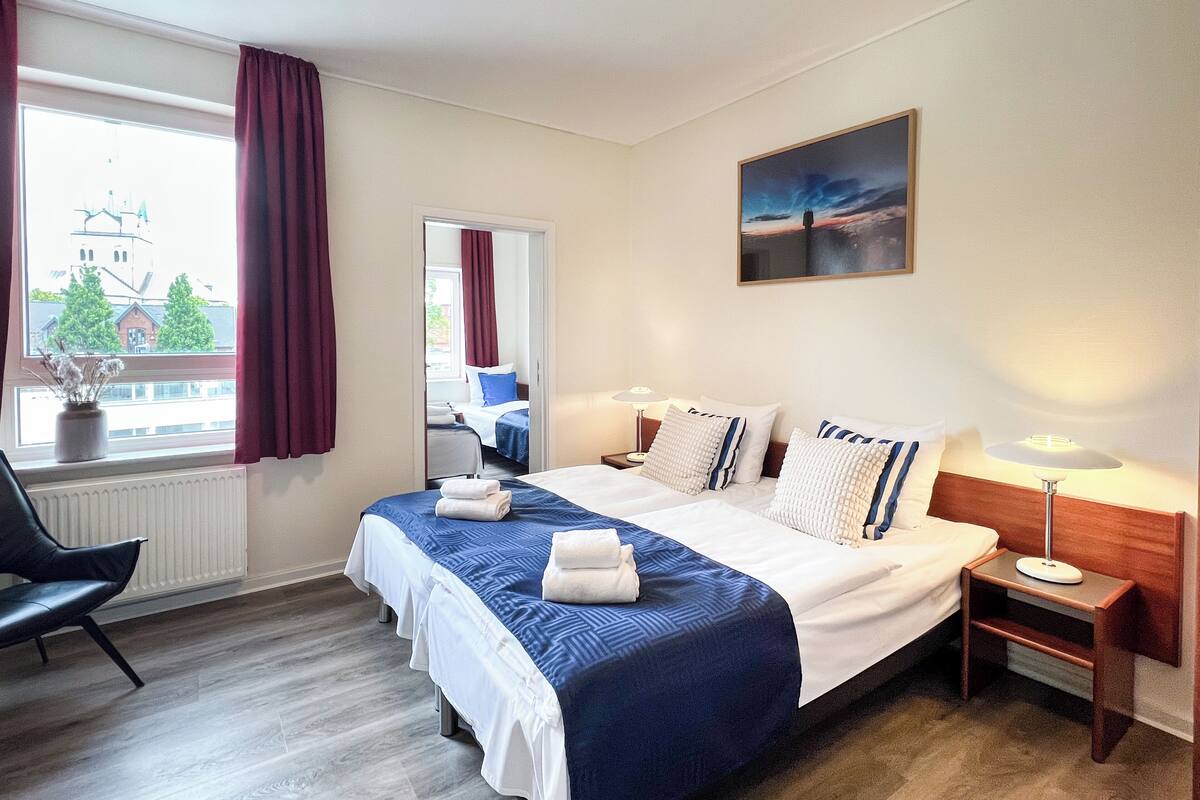 Quarto de hotel com uma cama grande, duas mesas de cabeceira, uma cadeira, uma janela com cortinas e vista para um edifício.