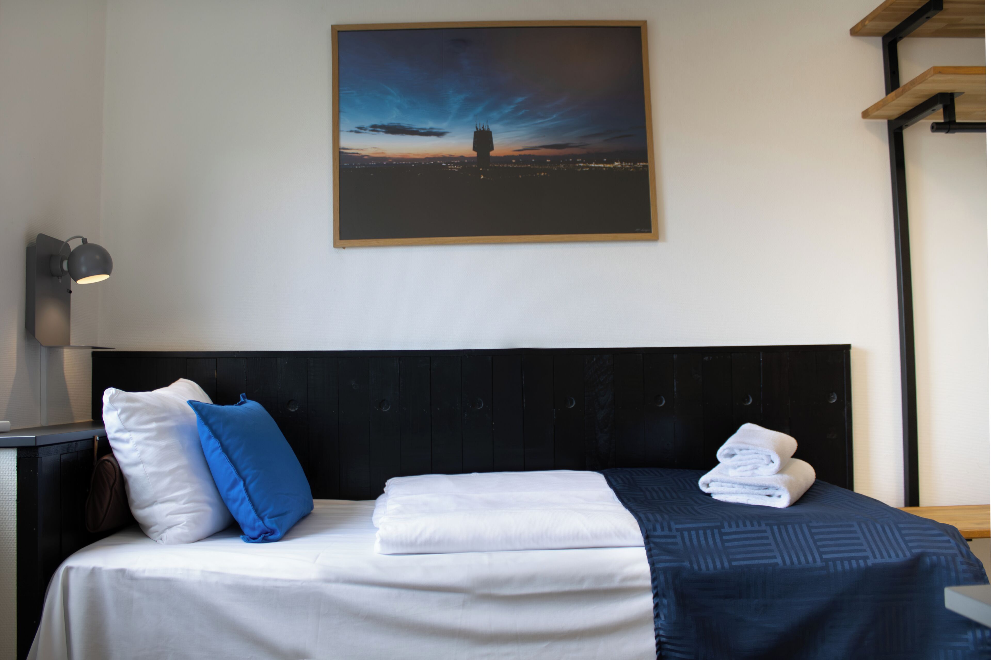 Ein ordentlich bezogenes Bett mit weißer und blauer Bettwäsche, ein gerahmtes Stadtbild im Abenddämmerung und eine moderne Nachttischlampe.