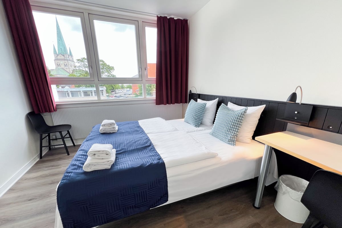 Quarto de hotel com cama, escrivaninha, cadeira e janela com cortinas.