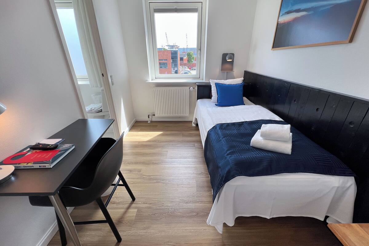 Quarto de hotel com cama, escrivaninha, cadeira, janela e um quadro na parede.