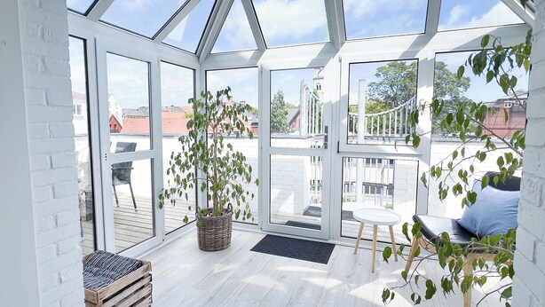 Amplia habitación con ventanas grandes, suelo de madera, una cesta de mimbre, un banco y una pequeña mesa con cojín.
