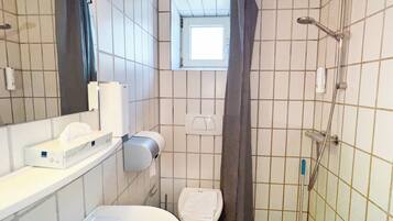 Une petite salle de bain avec des toilettes, un lavabo et une douche. Il y a une fenêtre au-dessus des toilettes et un distributeur de serviettes fixé au mur.