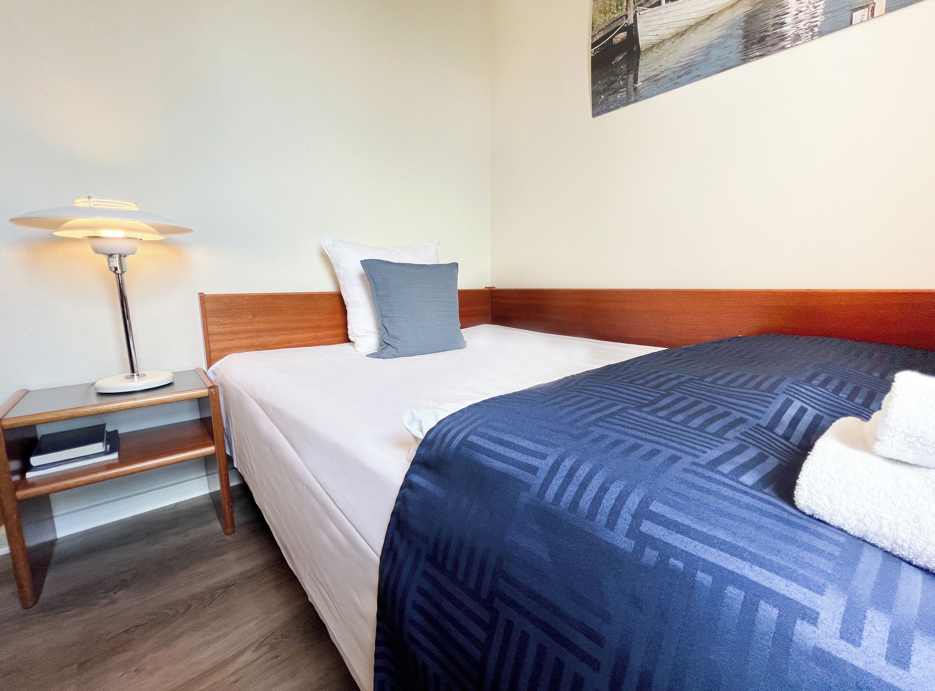 A neatly made bed with a blue patterned blanket, a wooden bedside table with a lamp, and a white pillow.