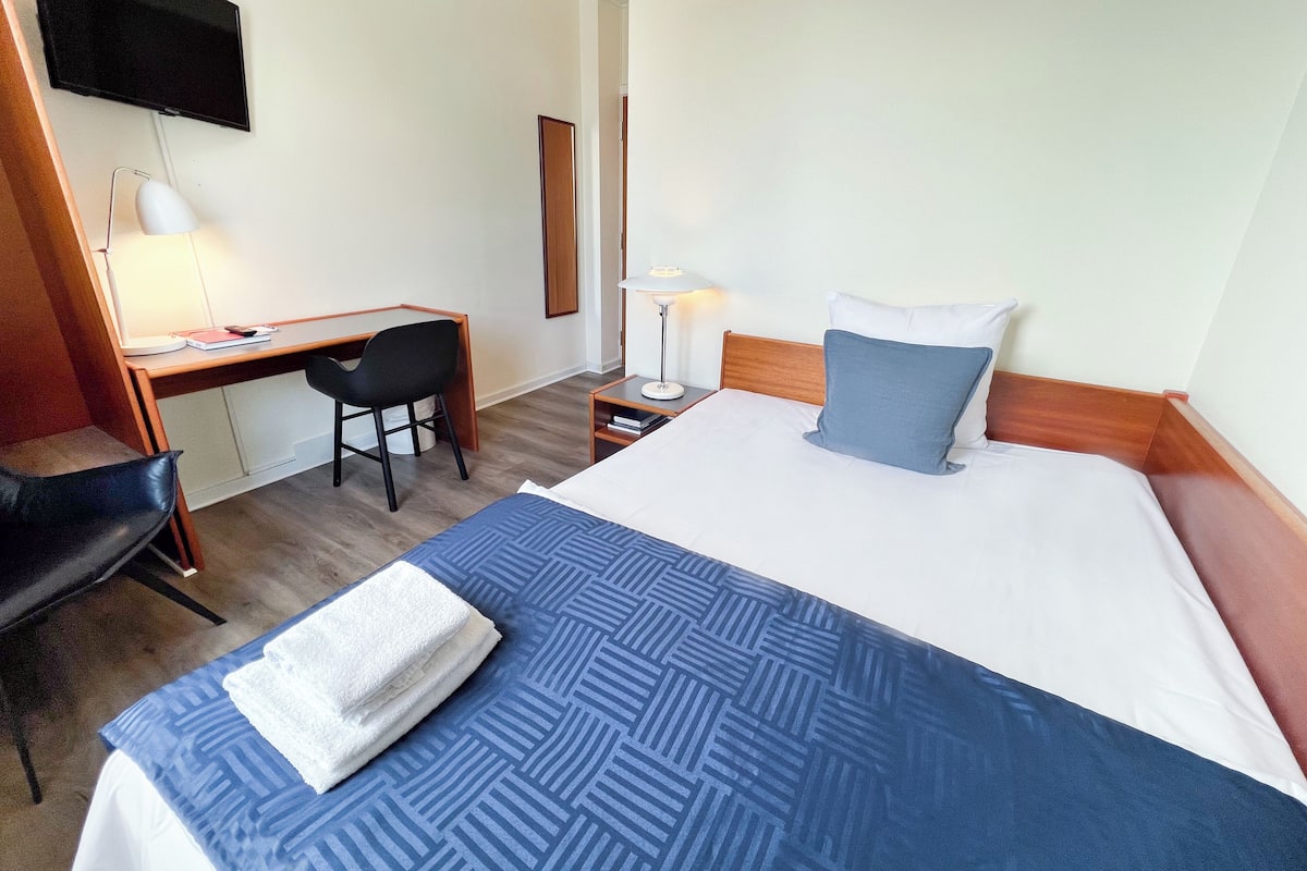 Quarto de hotel com cama, escrivaninha, cadeira, televisão e mesa de cabeceira com abajur.