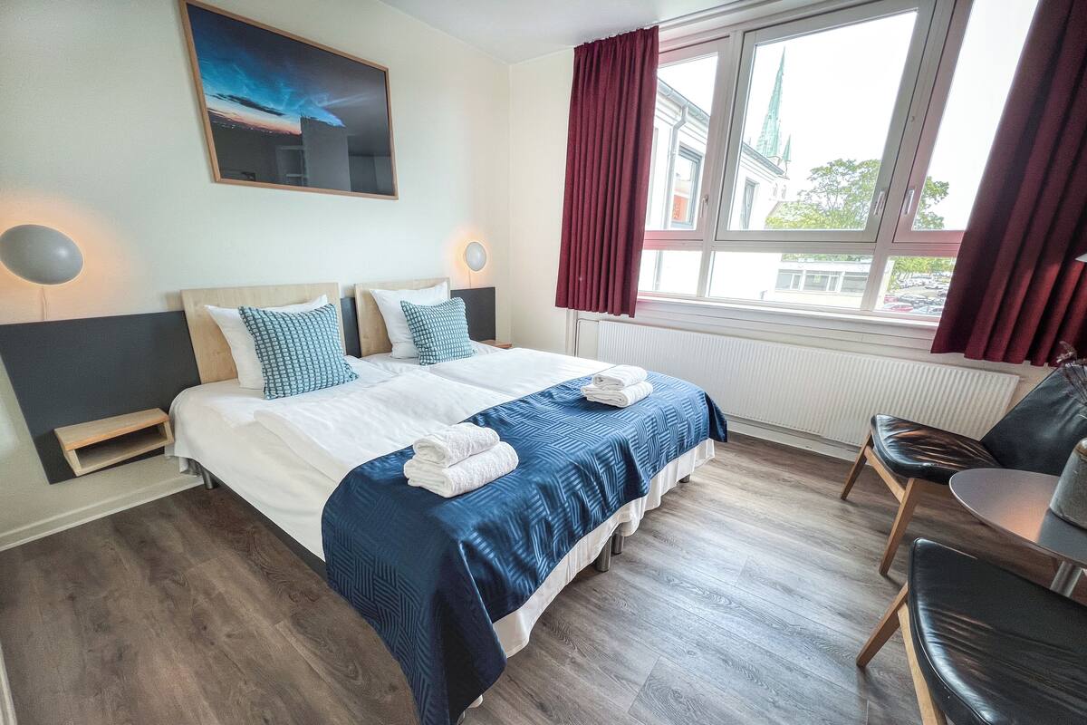 Quarto de hotel com uma cama grande, duas mesas de cabeceira, uma cadeira, janela com cortinas e um quadro na parede.