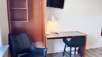 Een kleine, moderne kamer met een houten bureau, twee zwarte stoelen, een aan de muur bevestigde televisie en een lamp.