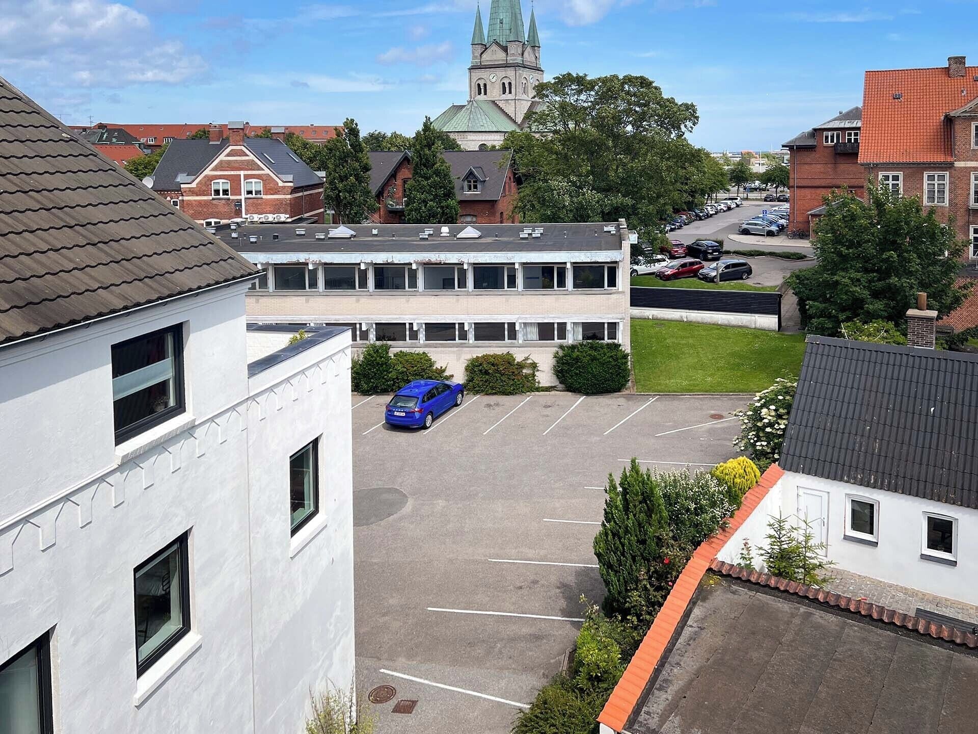 Un parking avec une voiture bleue, une église avec un clocher vert et des immeubles d’habitation.