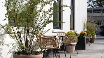 A patio with wicker chairs and potted plants, a white building with large windows, and a clear sky.