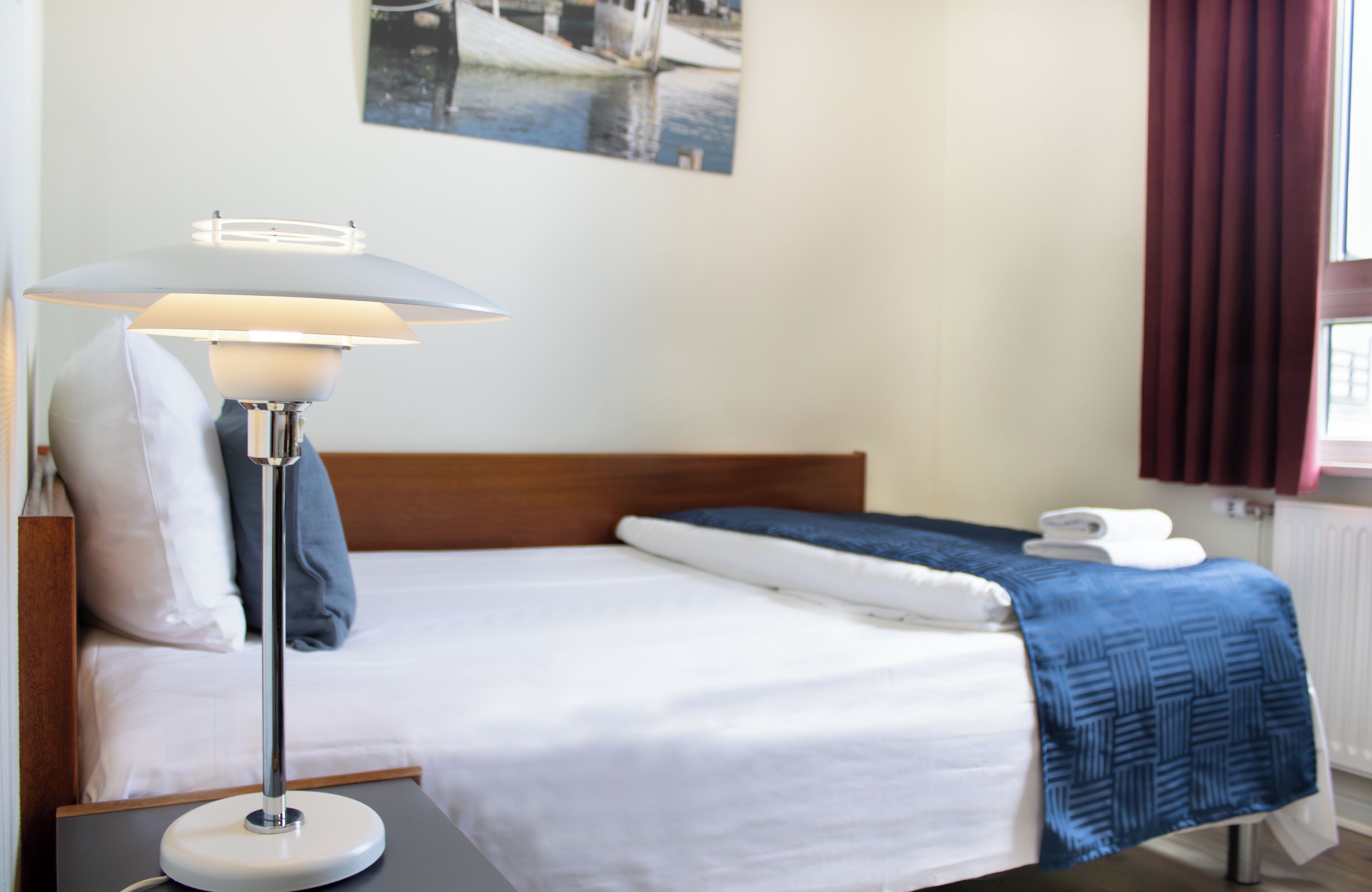 Un lit bien fait, avec du linge de lit blanc, une couverture bleue et une lampe de sol dotée d’un abat-jour blanc.