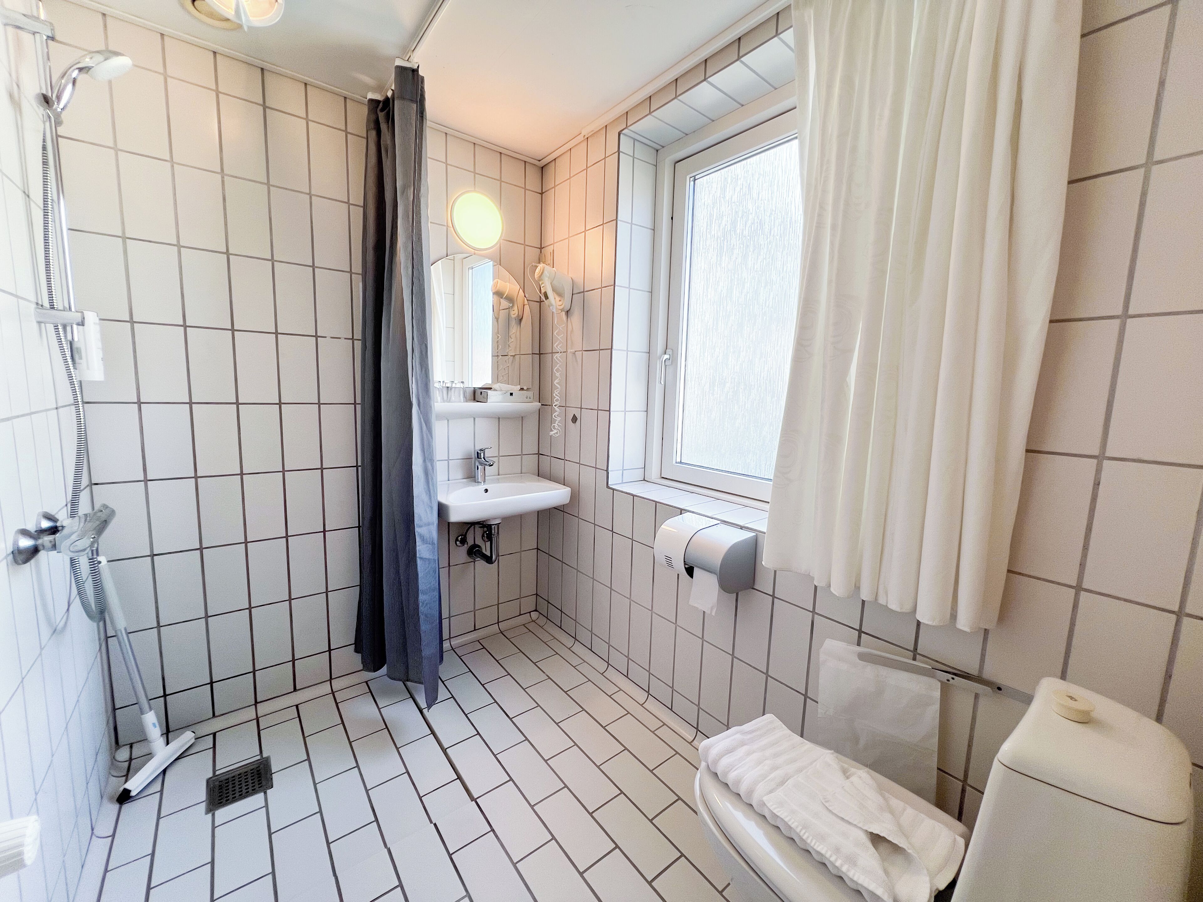 A bathroom with a shower, sink, toilet, and a window with curtains.
