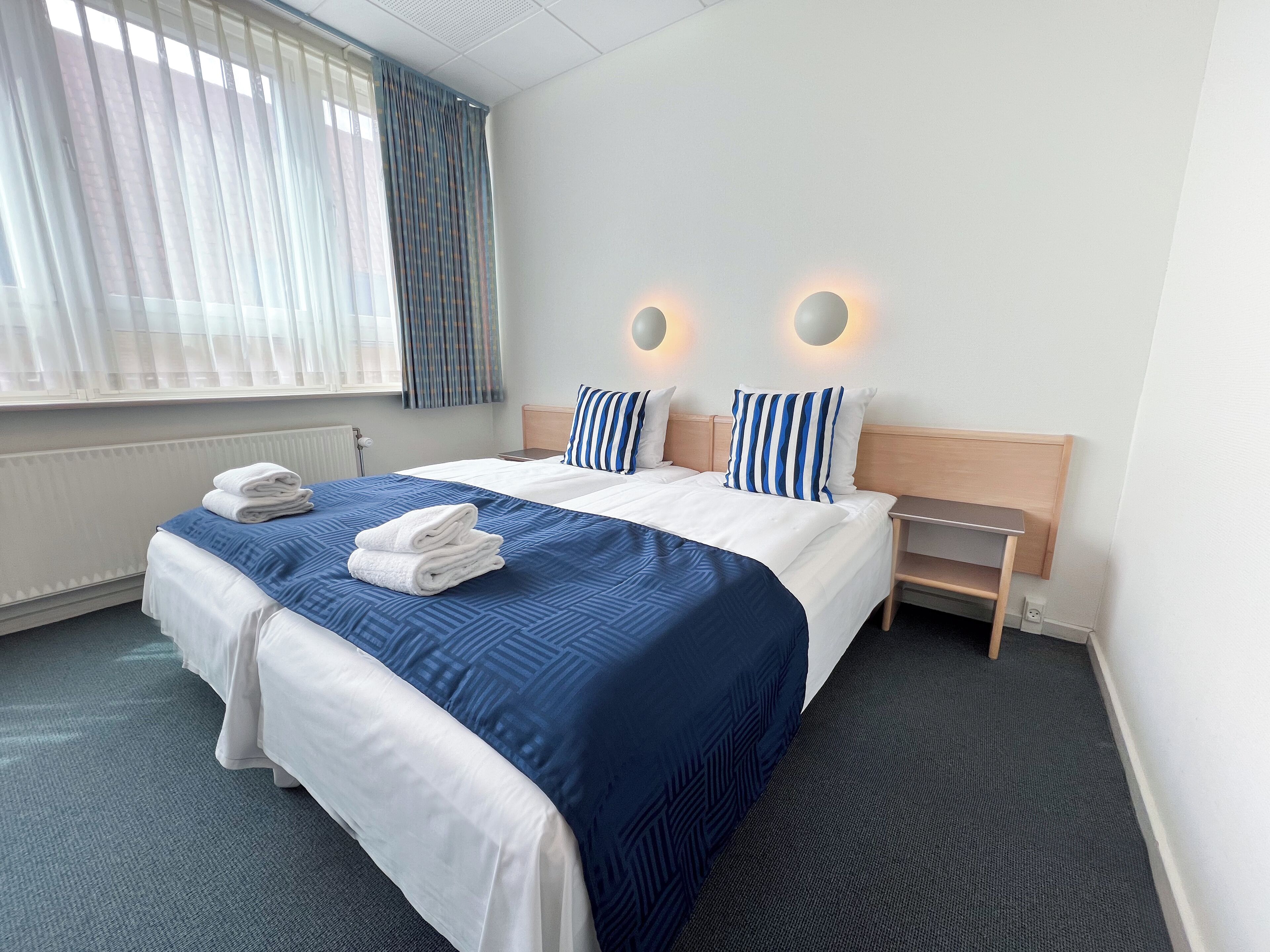 Ein Hotelzimmer mit einem großen Bett, zwei Nachttischen, einem Fenster mit Vorhängen und einer Wandleuchte.