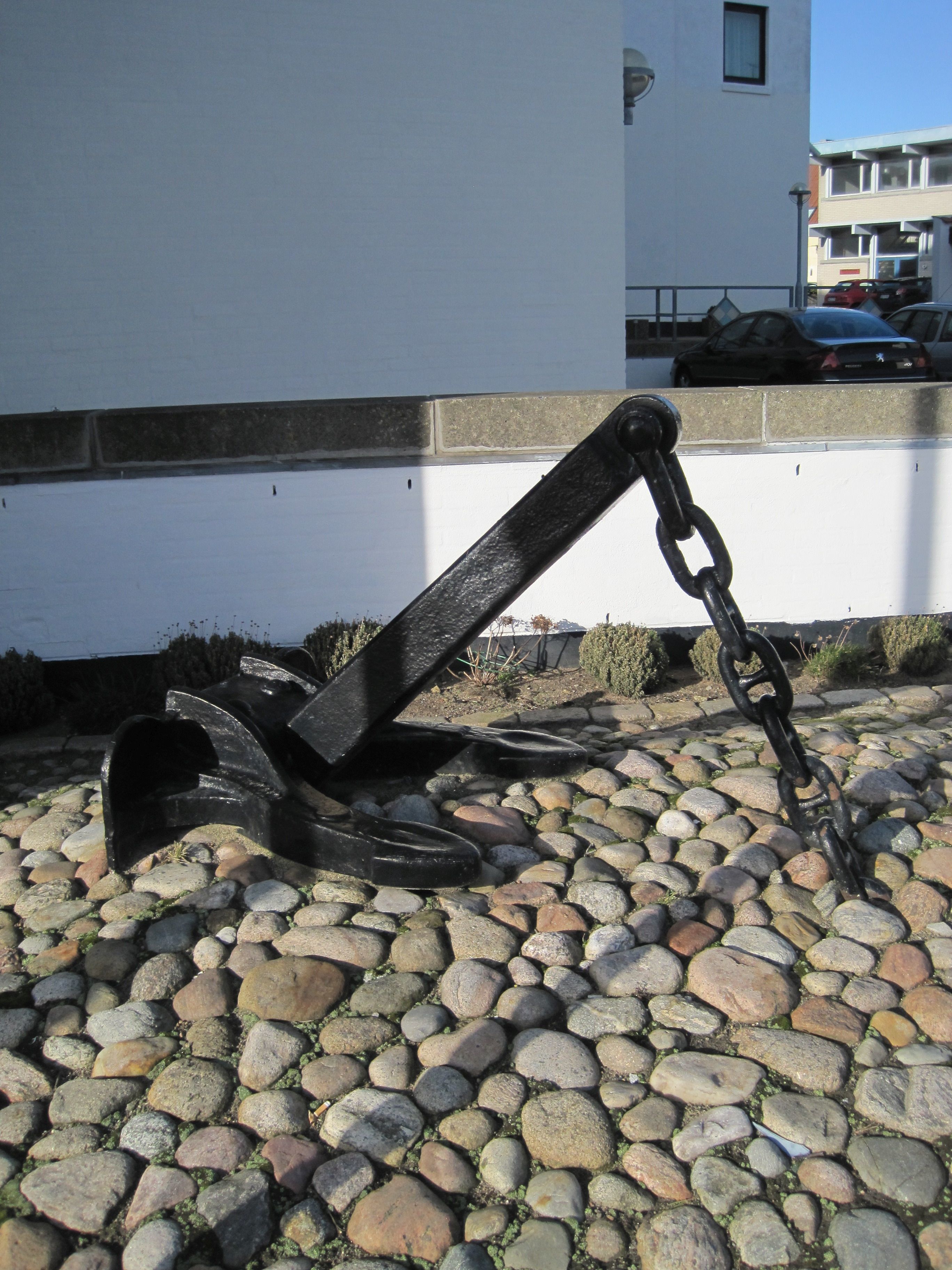 A black metal anchor on a stone surface with a white building in the background.