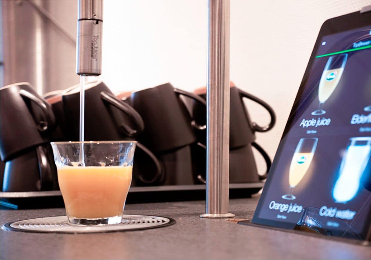 En juiceautomat med ett glas apelsinjuice på ett underlägg, och en digital meny som visar vilka juicer som finns att välja på.