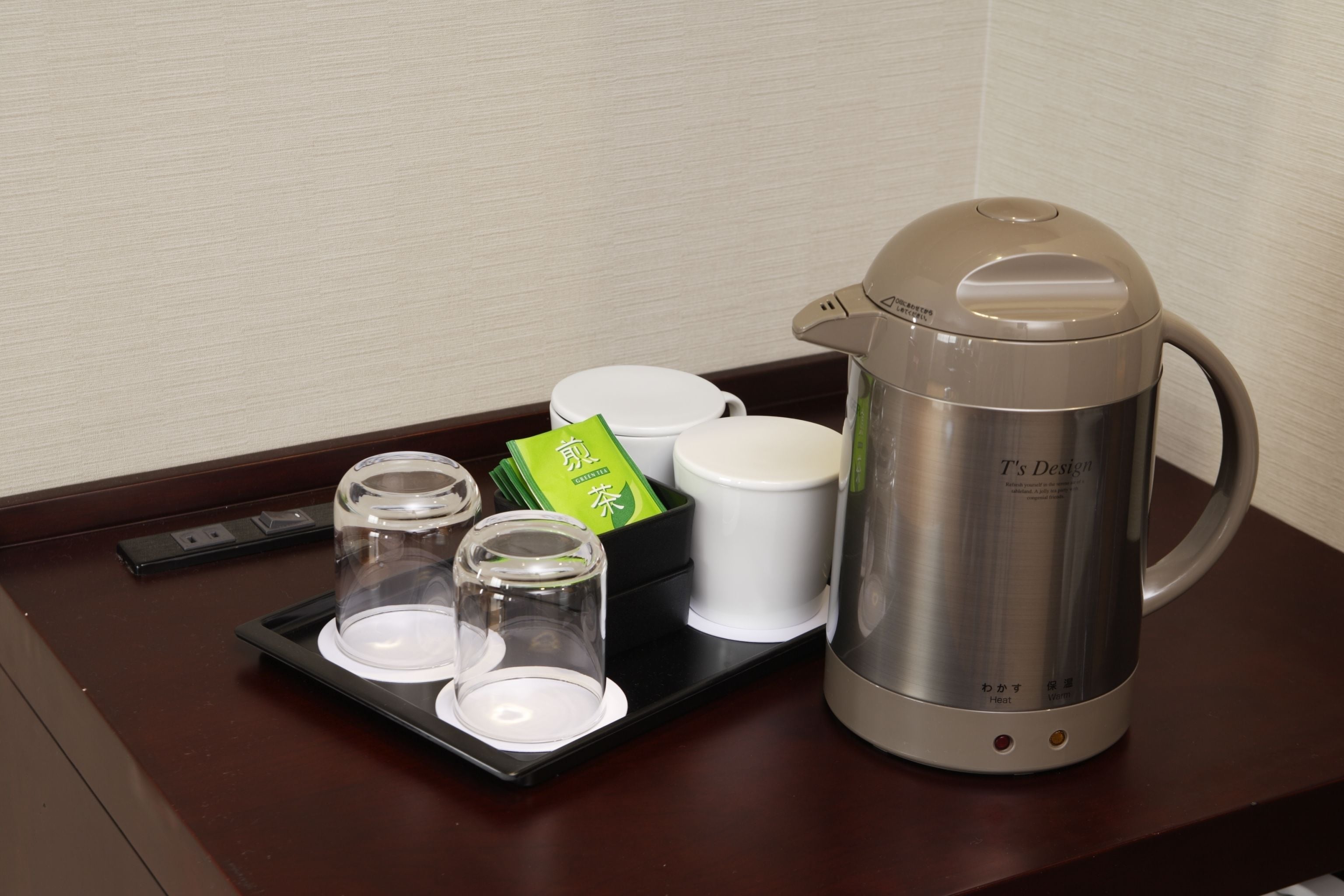 A stainless steel T's Design electric kettle on a tray with three glass cups and two white containers, likely for tea or coffee, on a wooden table.