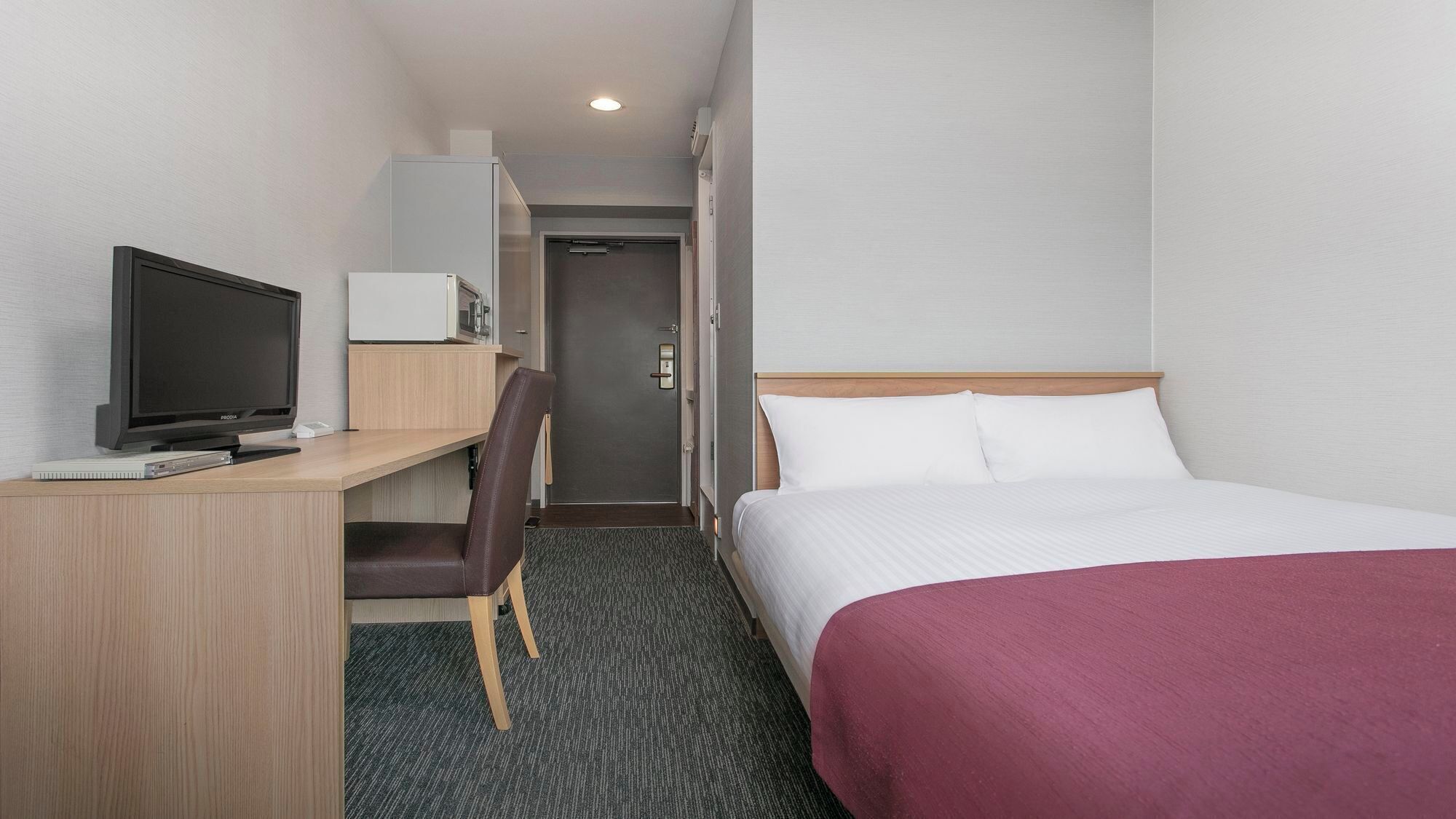 A hotel room with a bed, a desk with a television, a chair, and a door leading to a bathroom.