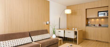 A modern living room with a brown sofa, a small table with candles, and a kitchen area with wooden cabinets and a microwave.