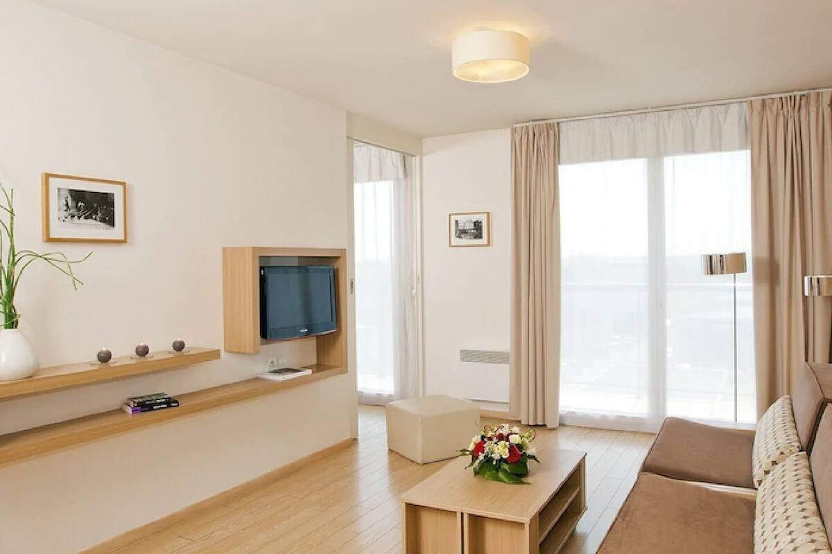 A modern hotel room with a wooden floor, a sofa, a TV on a wooden shelf, a small table with a vase of flowers, and a large window with curtains.