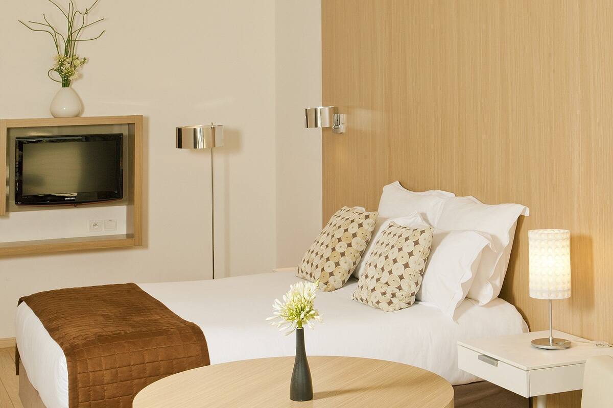 A modern hotel room with a bed, bedside table, lamp, TV, and a round table with chairs.