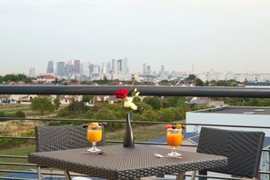 A balcony with a table set for two, featuring a vase with flowers and two drinks. A city skyline is visible in the background. - Residhome Carrières La Défense (Carrieres-sur-Seine)