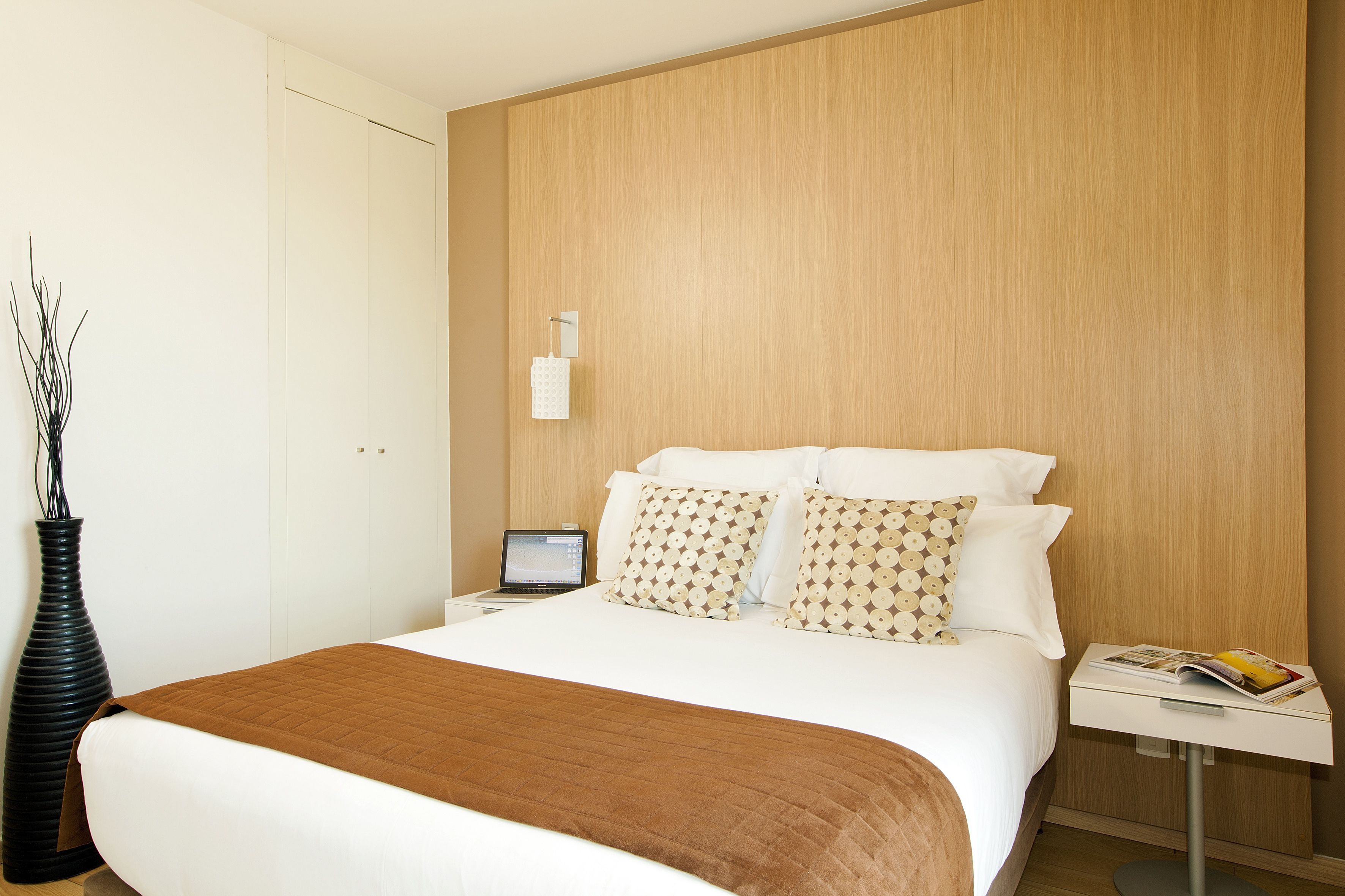 A neatly made bed with a brown blanket, a laptop on a bedside table, and a magazine on a nightstand.