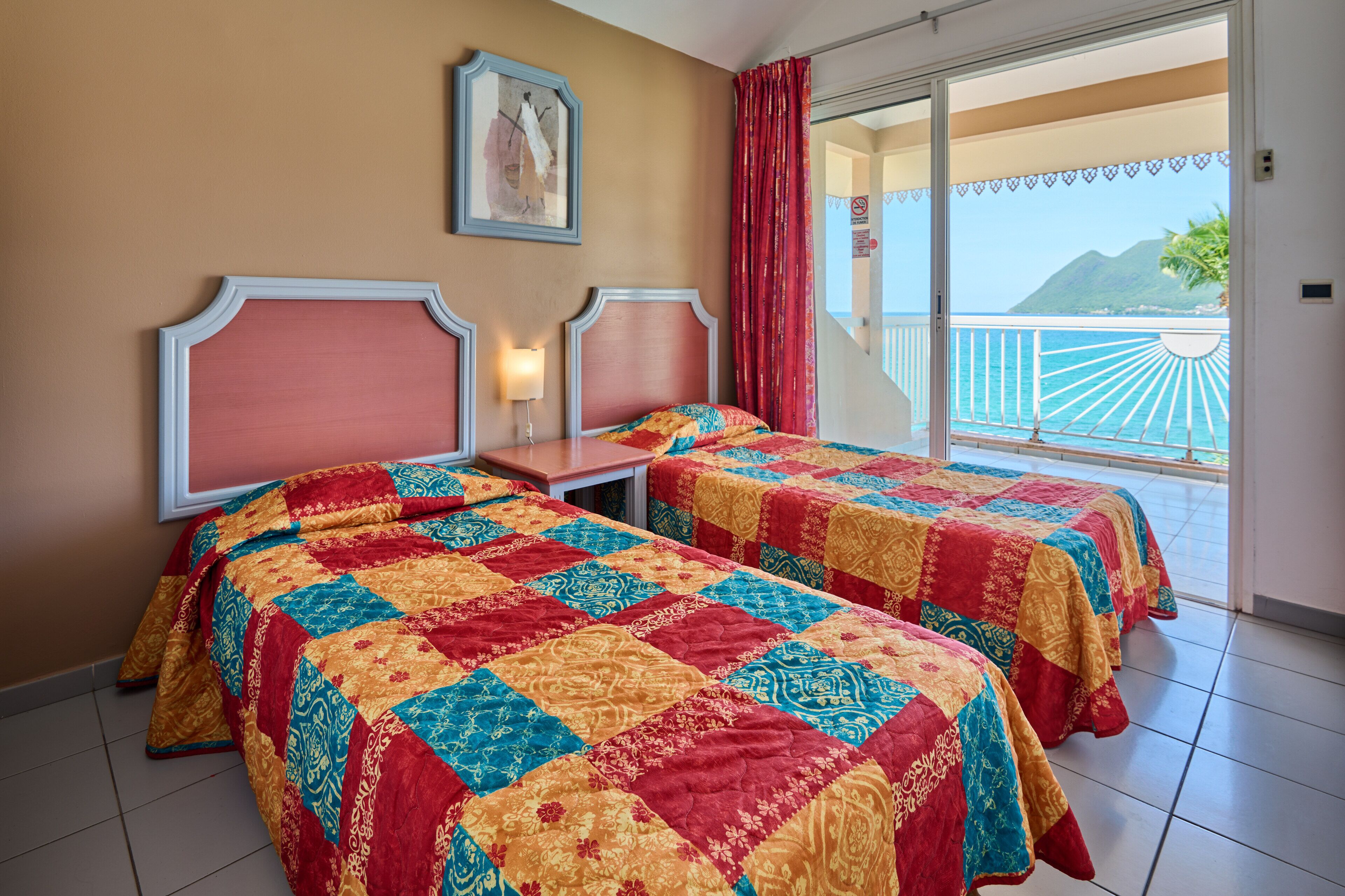 A hotel room with two beds, a nightstand, a painting on the wall, and a balcony with a view of the sea and mountains.
