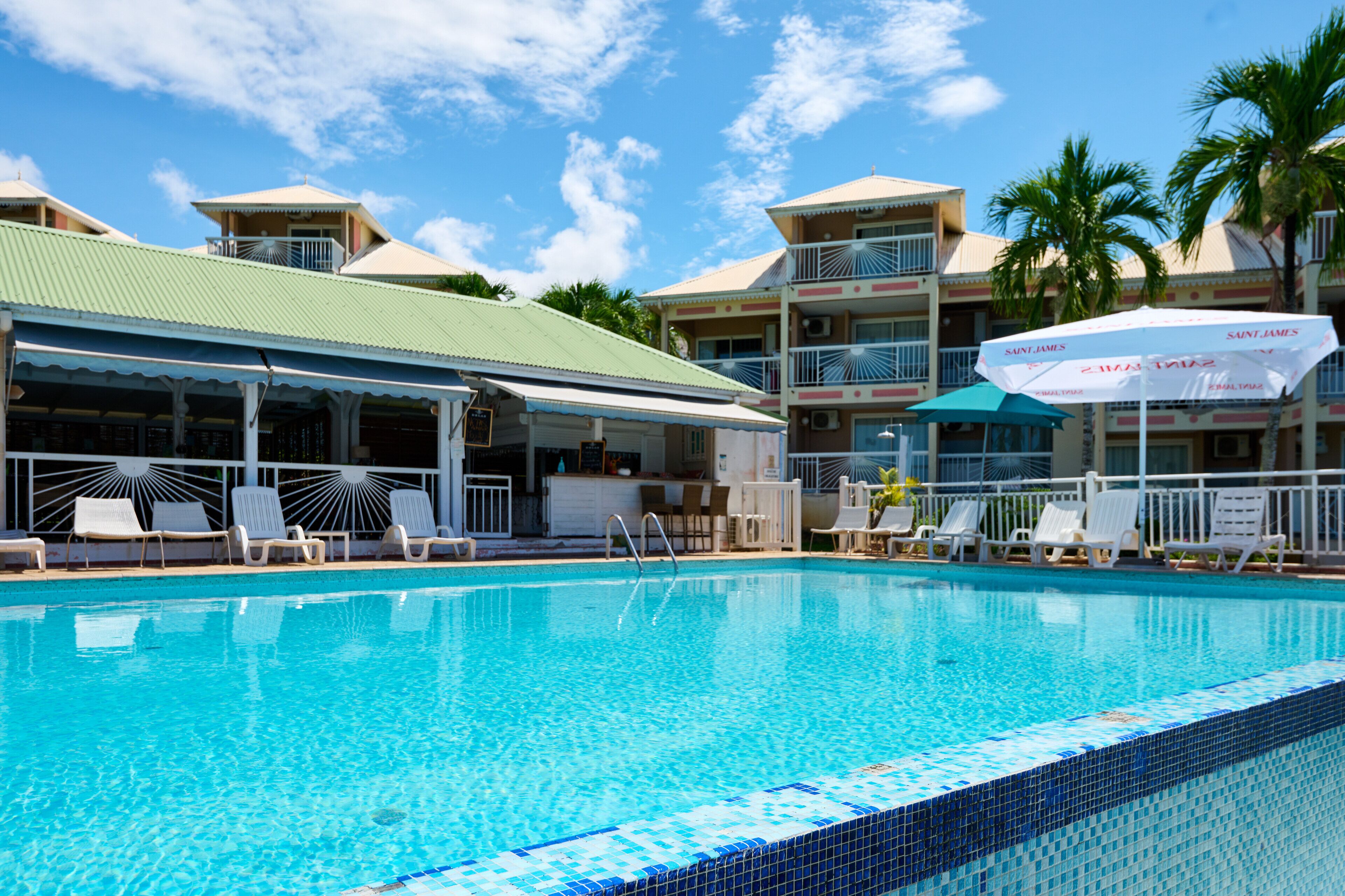 A swimming pool with lounge chairs and umbrellas, surrounded by multi-story buildings with balconies and palm trees.