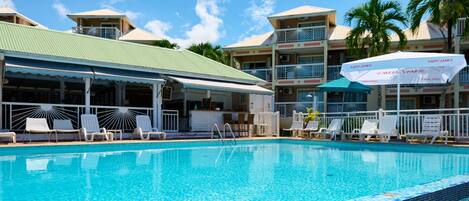 A swimming pool with lounge chairs and umbrellas, surrounded by multi-story buildings with balconies and palm trees.