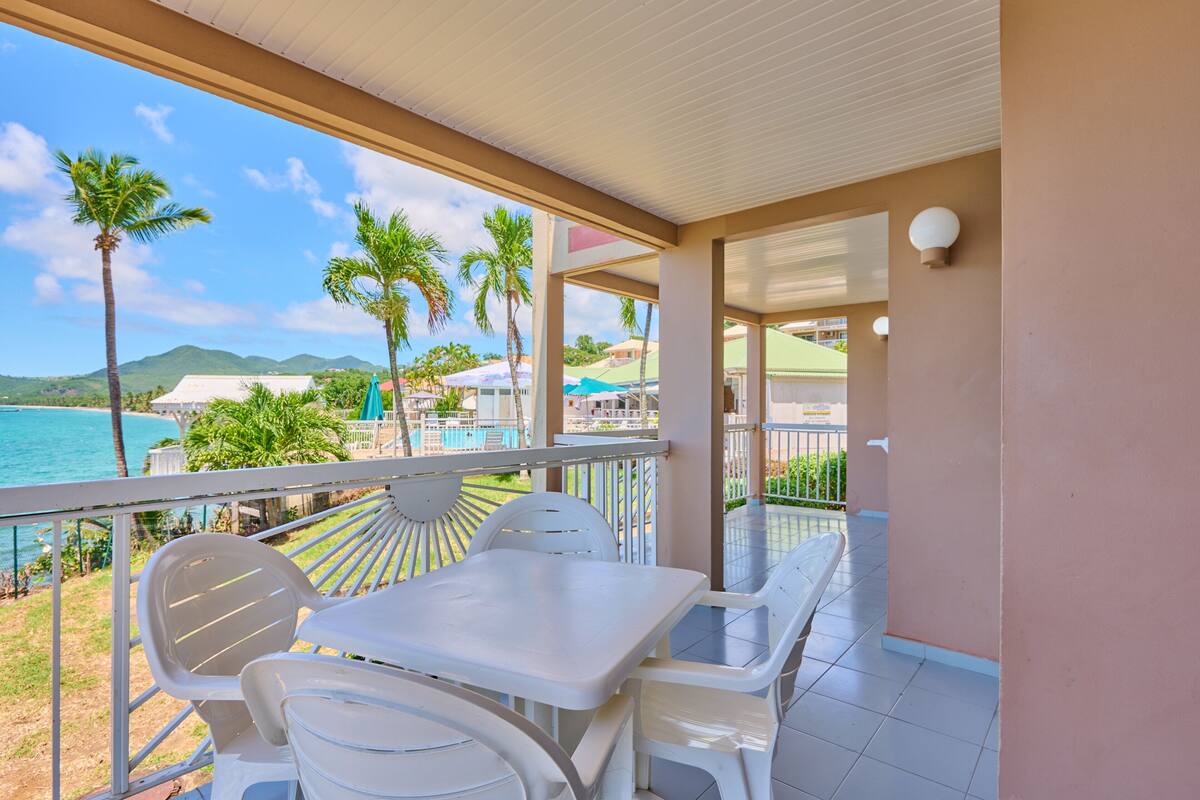 A balcony with white plastic chairs and a table, overlooking a pool and palm trees.