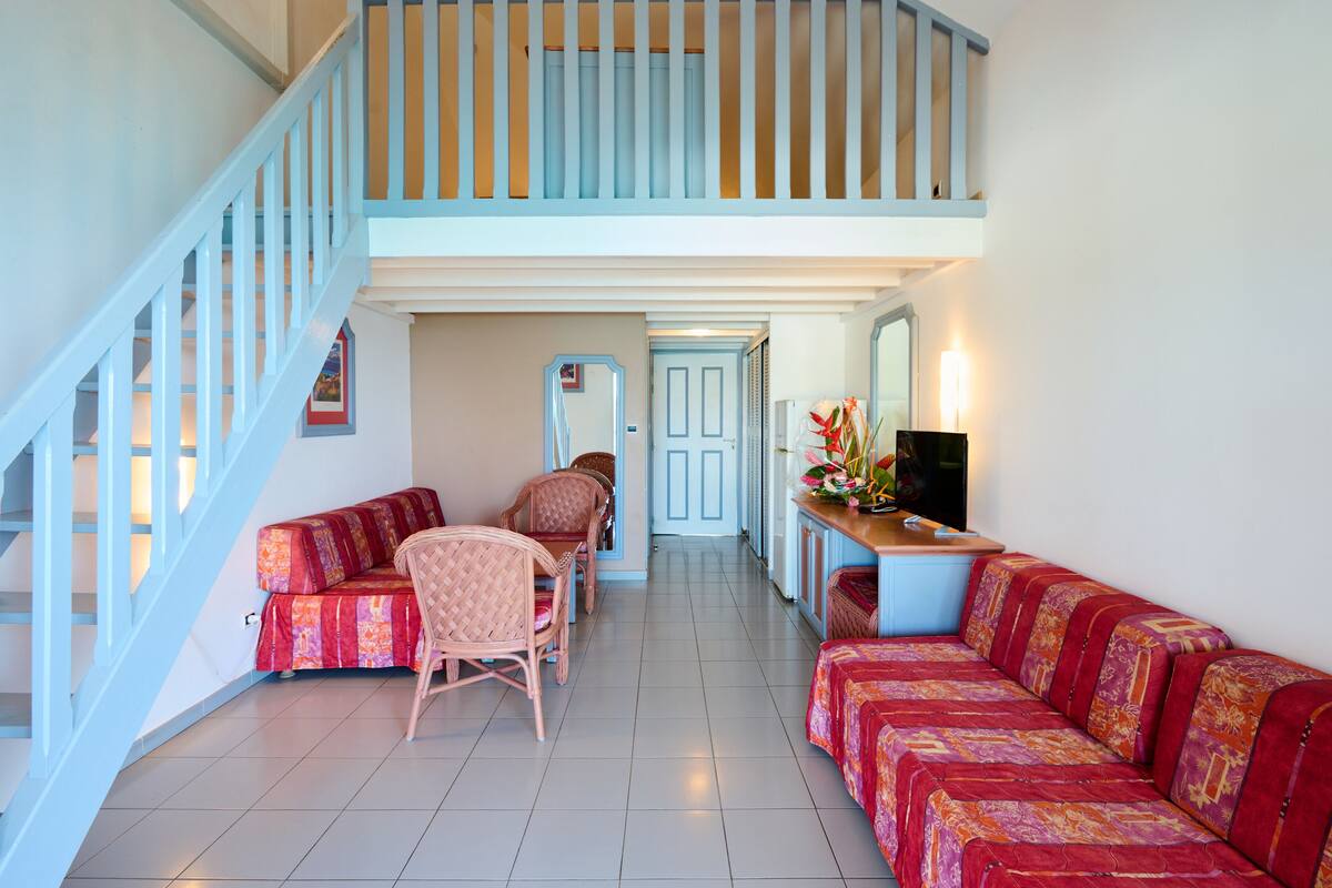 A spacious living area with a staircase, a sofa set, a dining table with chairs, and a television on a stand.