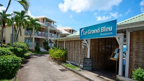 A sign for Le Grand Bleu Restaurant, a building with balconies, and a pathway lined with greenery. - Diamant Beach (Le Diamant)