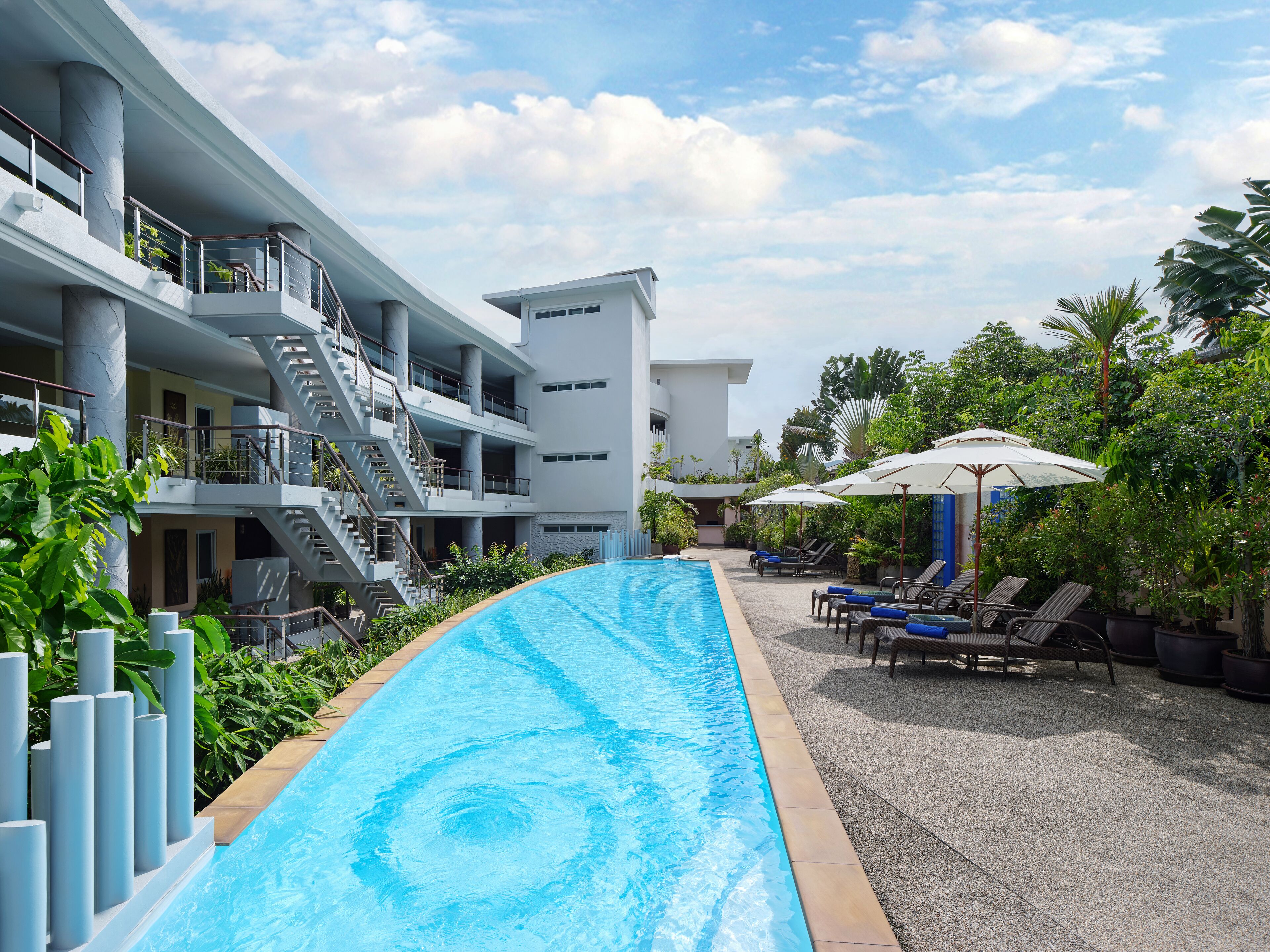 A swimming pool with a modern building in the background, surrounded by greenery and lounge chairs.