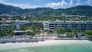 A beachfront hotel with multiple floors, palm trees, and a view of the beach and mountains.