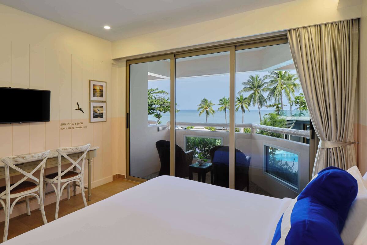 Habitación de hotel con cama, televisor, escritorio con sillas y balcón con vista a la playa y palmeras.