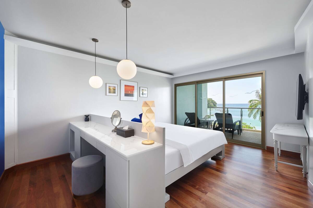 Una habitación de hotel moderna con una cama grande, un escritorio con espejo, una mesita y un balcón con vistas al océano.