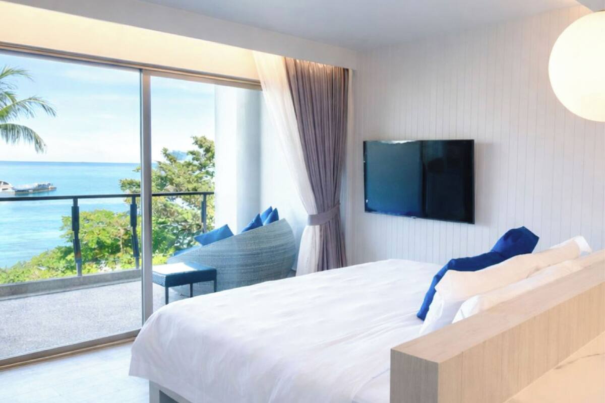 Habitación de hotel con una cama grande, un televisor montado en la pared y un balcón con vista al mar.