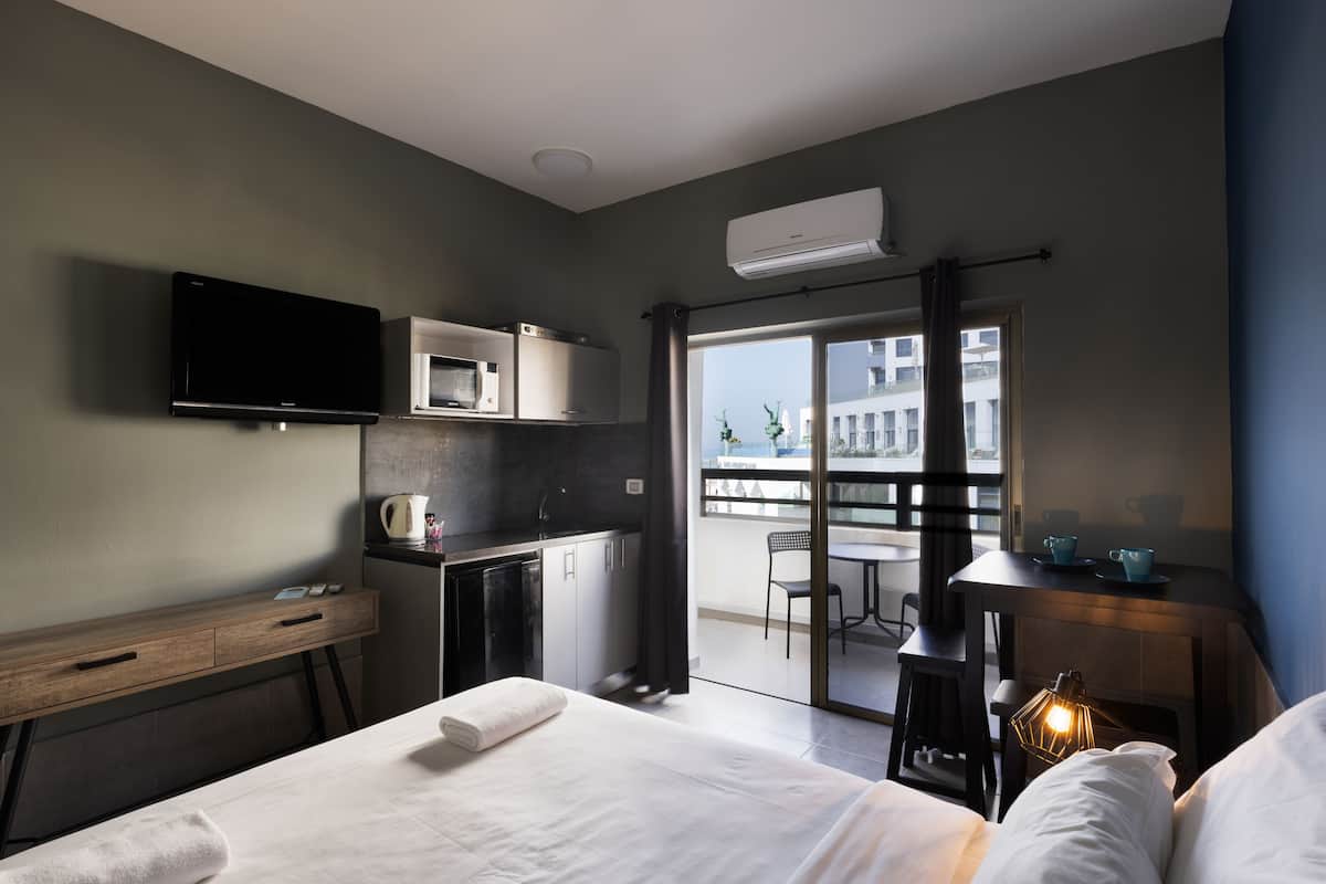 A hotel room with a bed, a desk with a lamp, a kitchenette, a TV, and a balcony with a view of buildings.