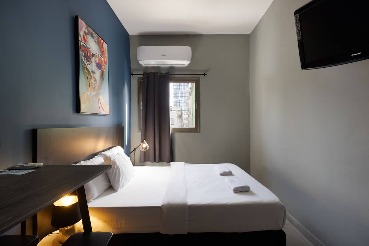 A hotel room with a bed, a desk, a lamp, a TV, and an air conditioner.