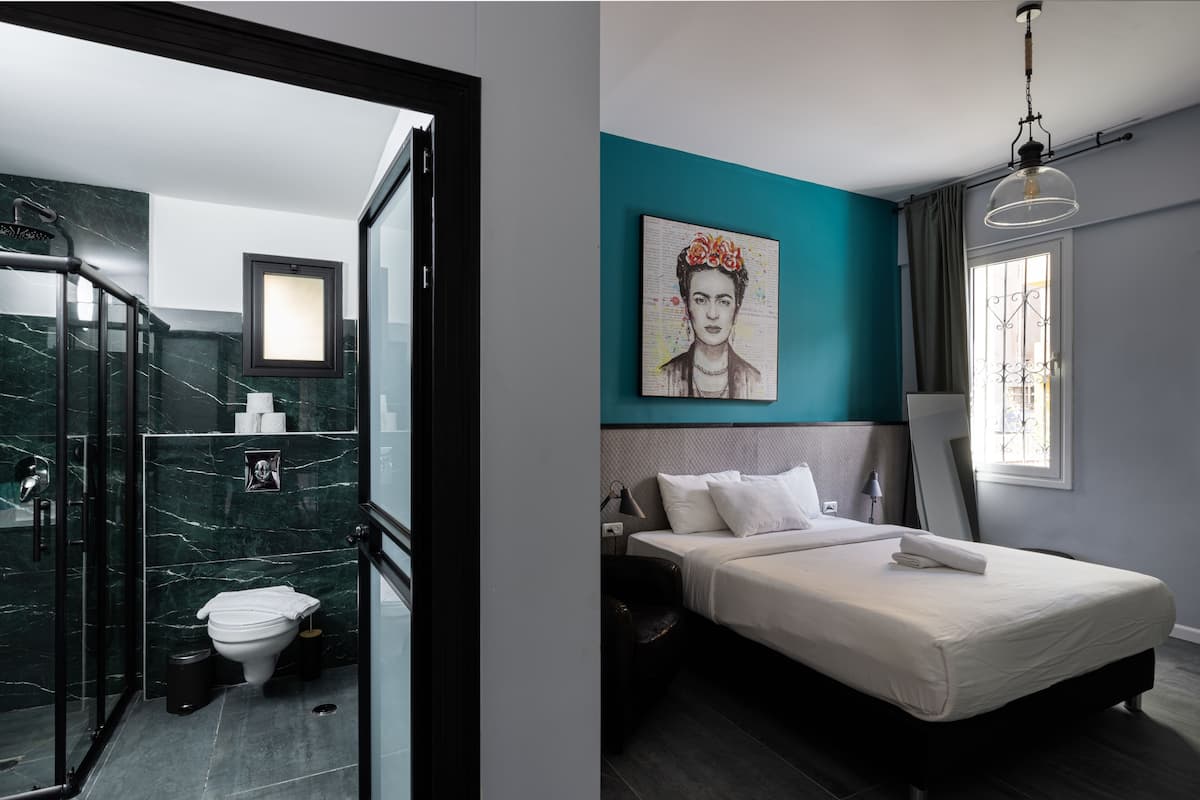 A modern bathroom with a glass shower enclosure, a toilet, and a marble wall. The bedroom features a large bed, a painting on the wall, and a window with curtains.