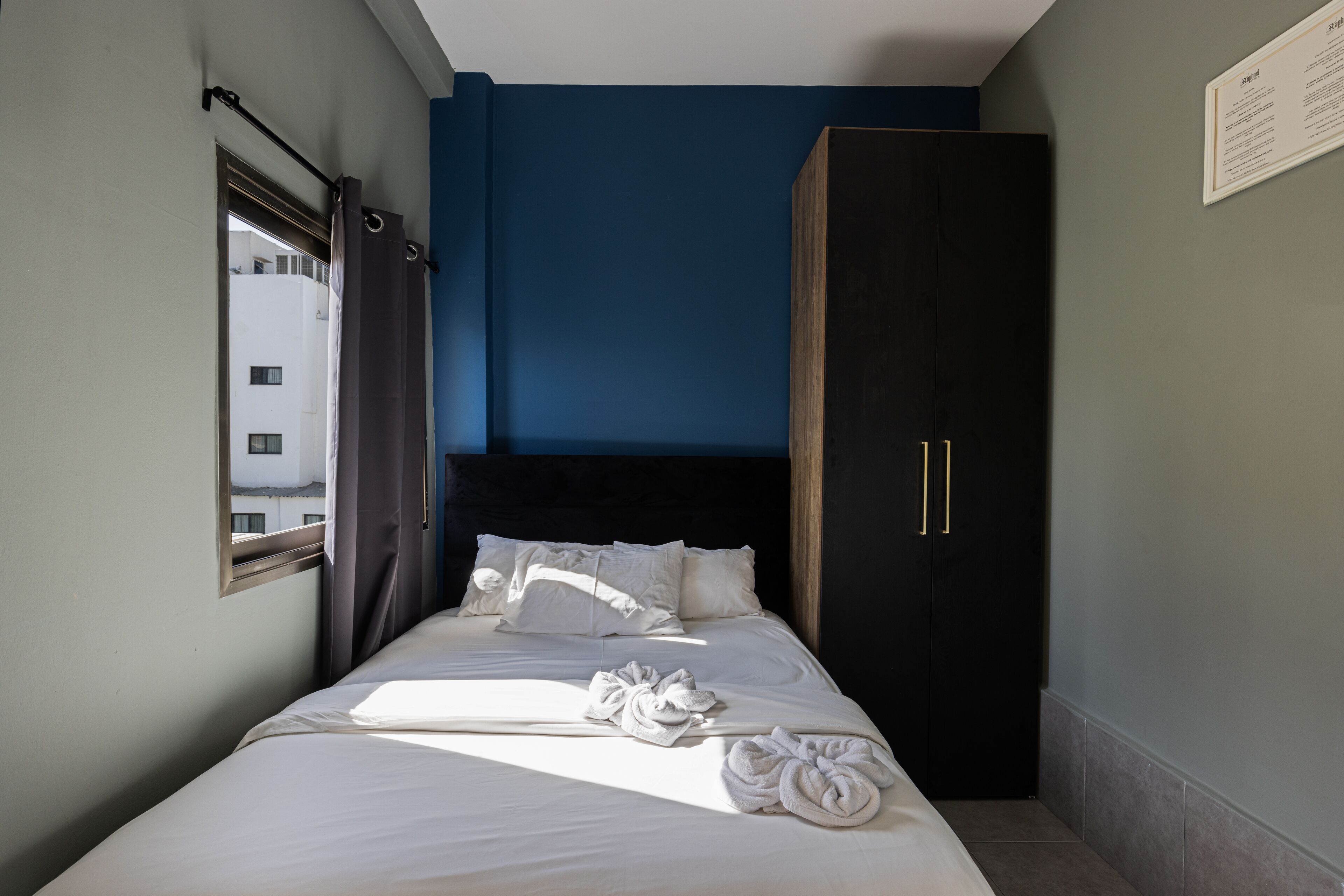 A hotel room with a bed, a wardrobe, a window with curtains, and a view of a building outside.