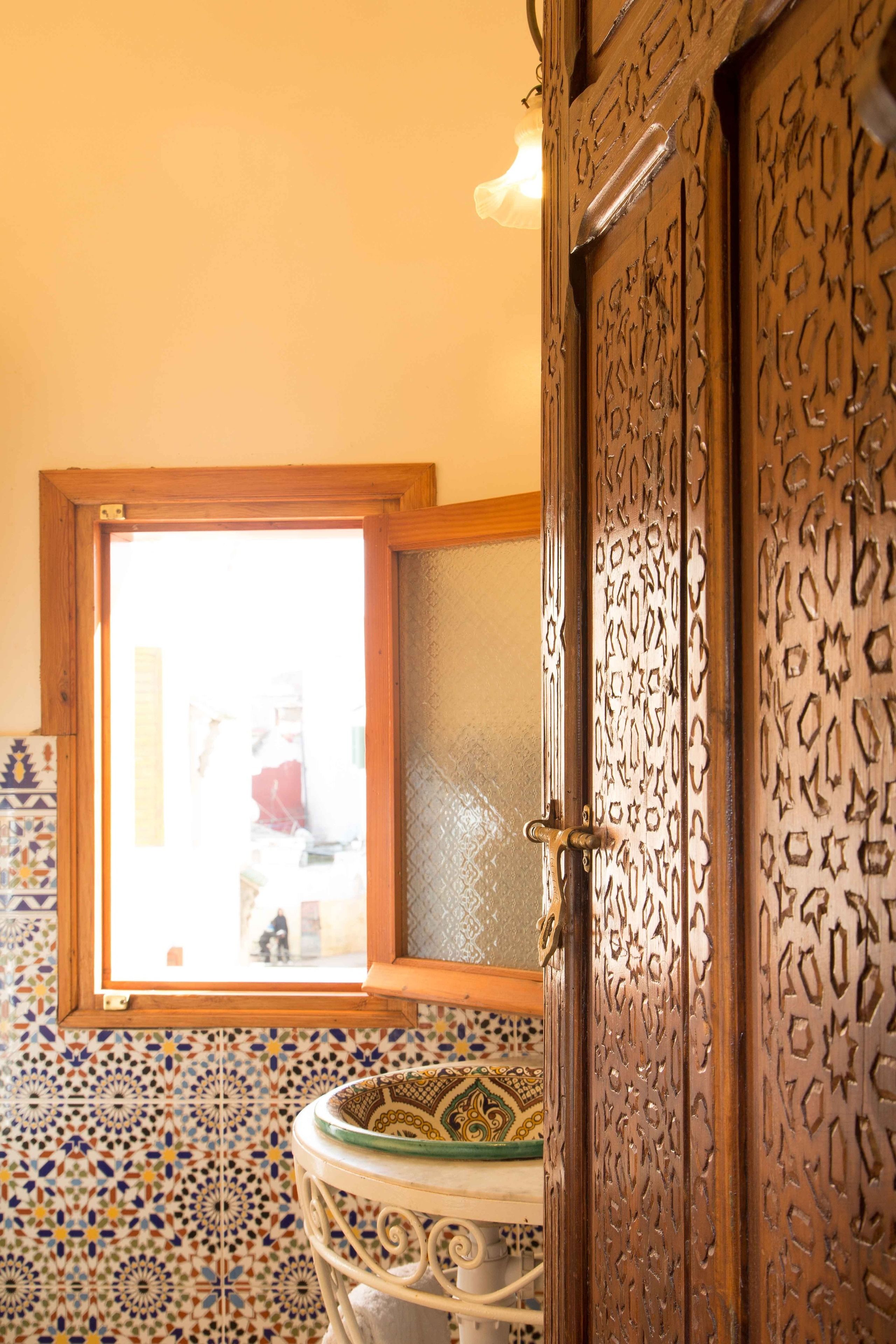 A bathroom with a patterned tile floor, a wooden door with intricate carvings, and a window with a view of the outside.
