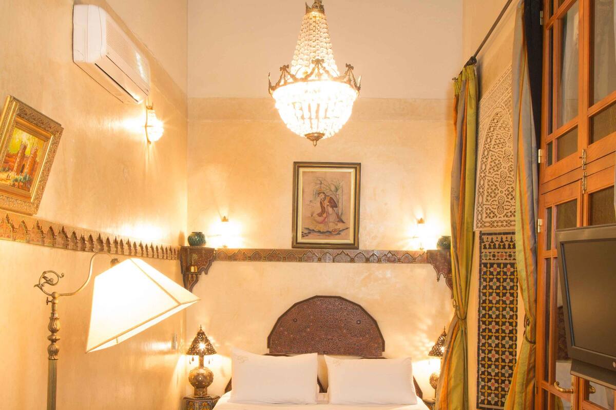 A room with a bed, a chandelier, a television, and a table with a vase of flowers.